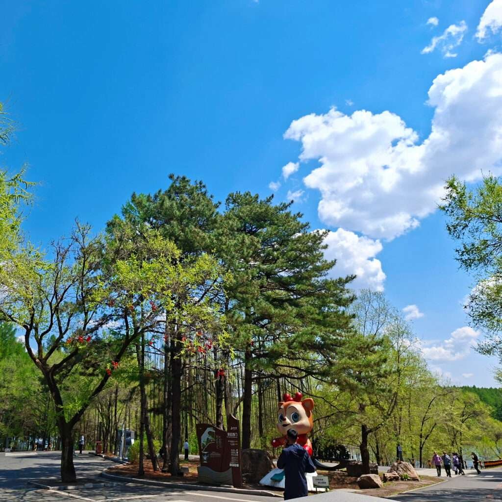 Park scene with blue sky, tall green trees, and a large cartoon squirrel statue near a walkway, with people strolling nearby.