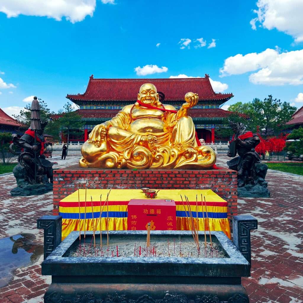 Gold Laughing Buddha statue seated on brick plinth in a temple courtyard with an incense burner in front and a blue sky overhead.