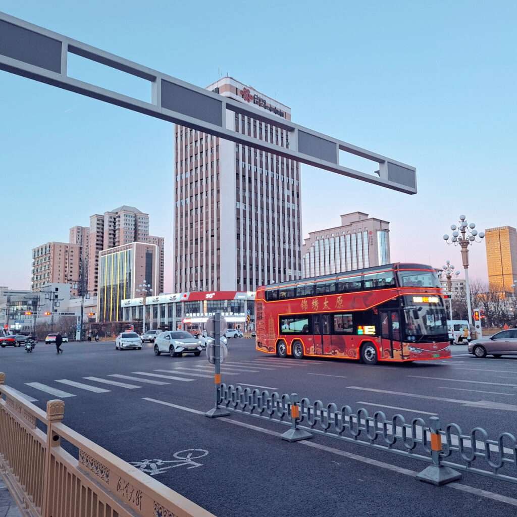 Red double-decker bus driving through a busy city intersection with tall office buildings in the background