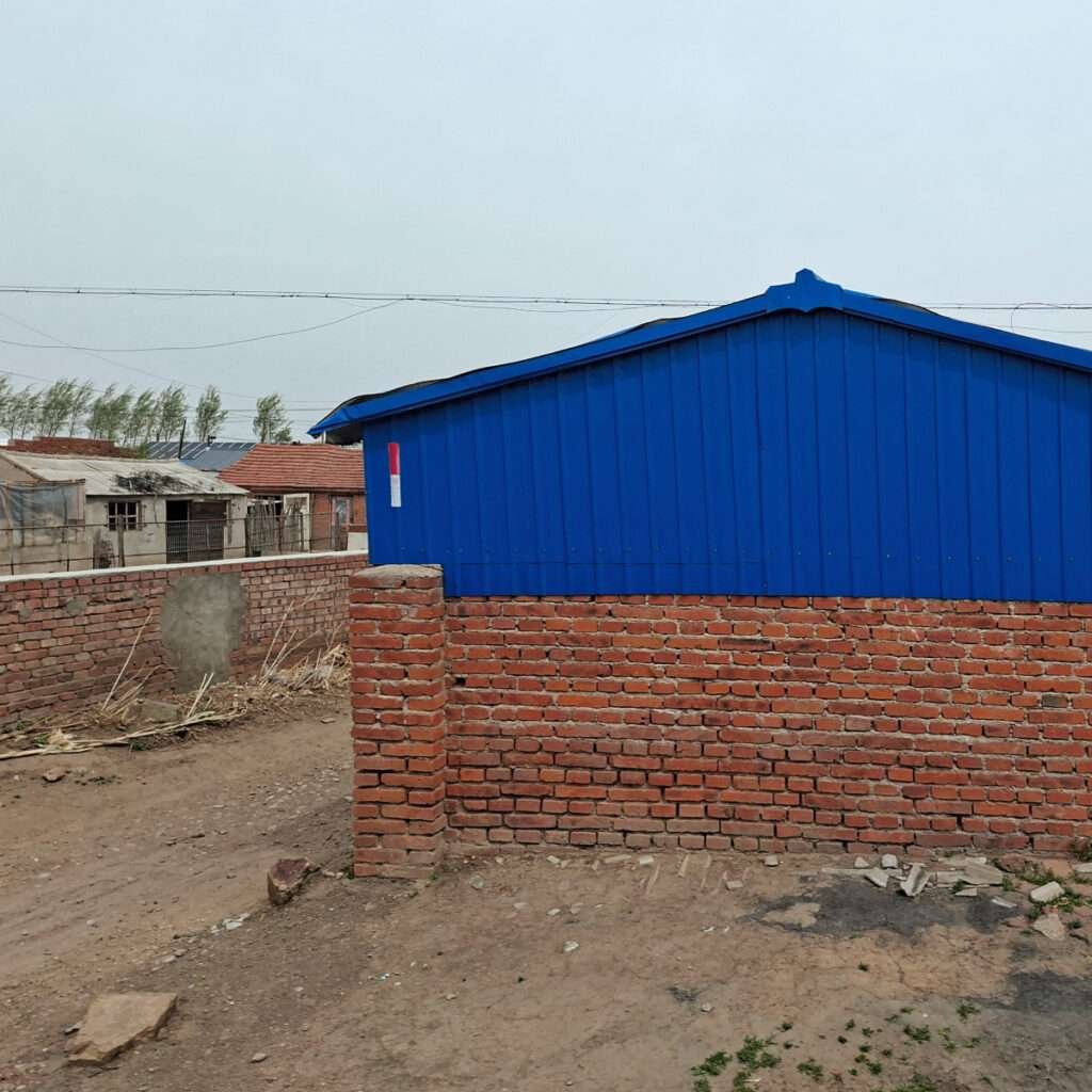 Bright blue corrugated metal building behind a low red brick wall on a dusty yard.