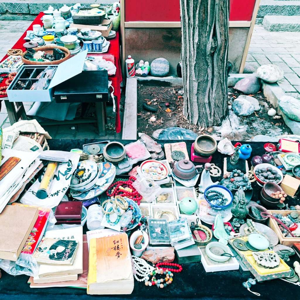Table of miscellaneous items at a street market: teaware, beads, stones, jewelry, books, and small sculptures.