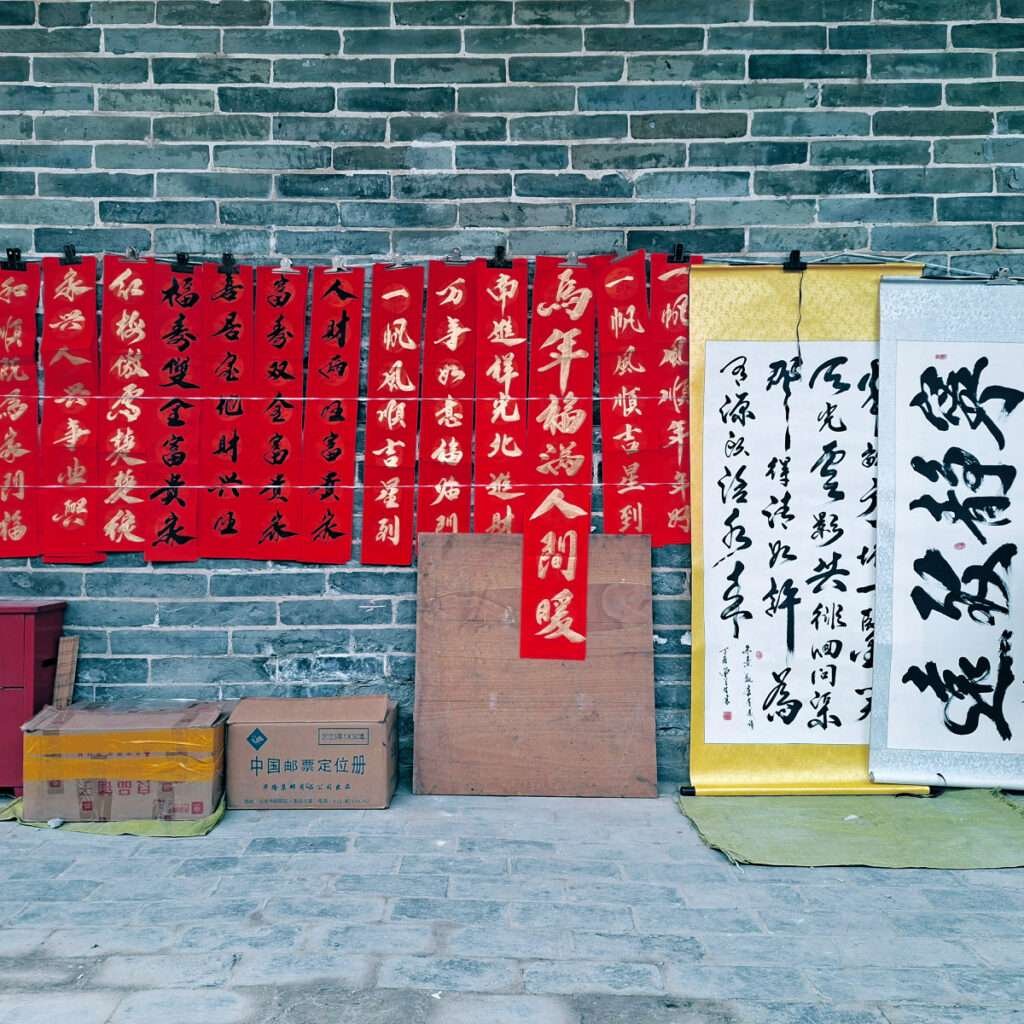 Red Chinese New Year banners with gold characters hung on a brick wall, with cardboard boxes on the ground.