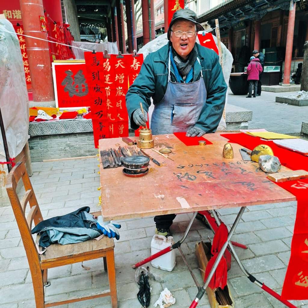 Elderly man in a cap and apron writes with a brush at a street stall selling Chinese calligraphy banners; red papers, ink pots, and brushes on a wooden table.
