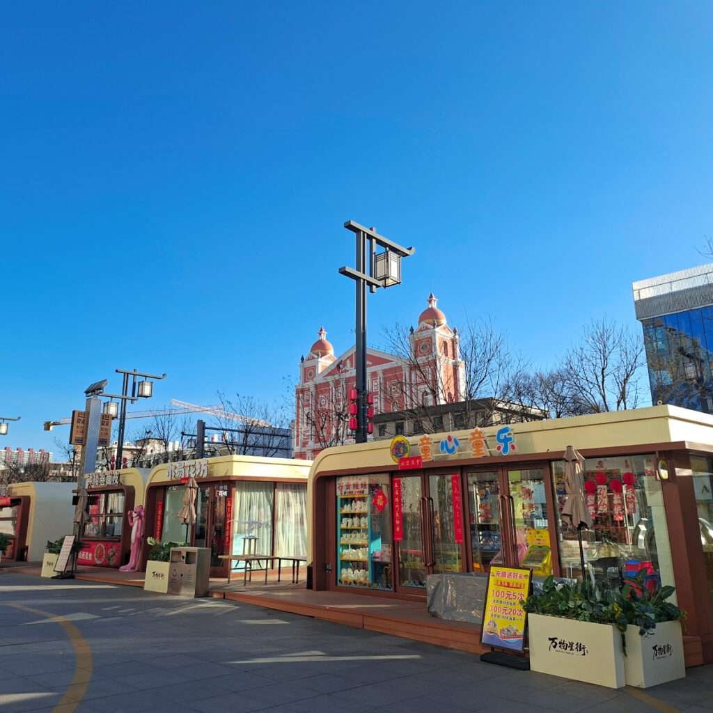 Sunny outdoor shopping street with pastel storefronts and Chinese signs; pink church-like building with domed towers in the background.