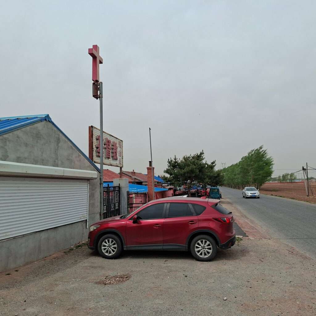 Red SUV parked on a gravel lot beside a storefront with a shuttered window and a Chinese sign on a pole.