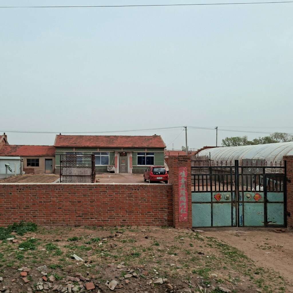 Brick wall and gate enclosing a rural courtyard with a red car parked near a green-roofed house; a white greenhouse on the right and a clear blue sky above.