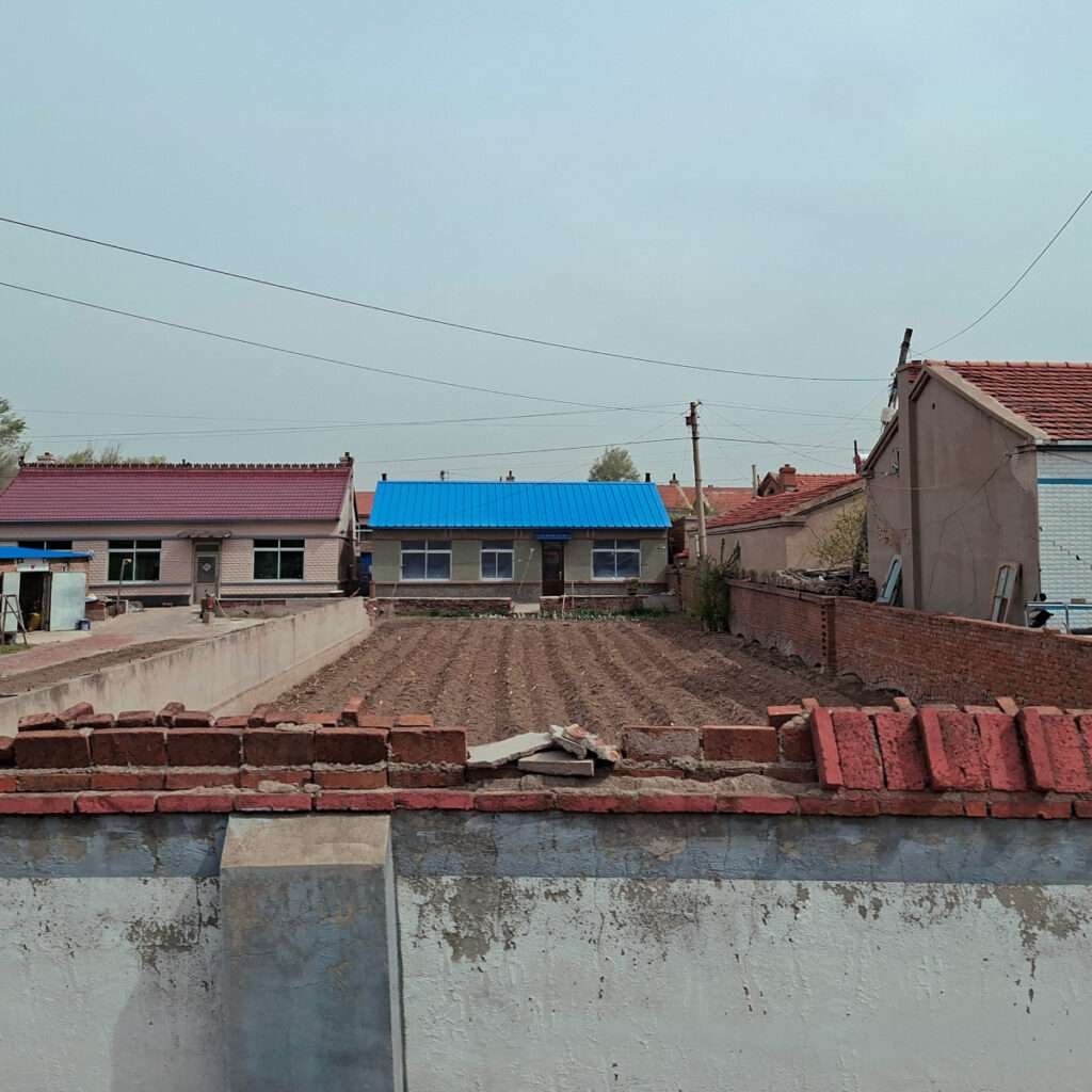 Row of small houses with red tile roofs and a blue metal-roofed building, behind a tilled field and a low brick wall under a pale sky.