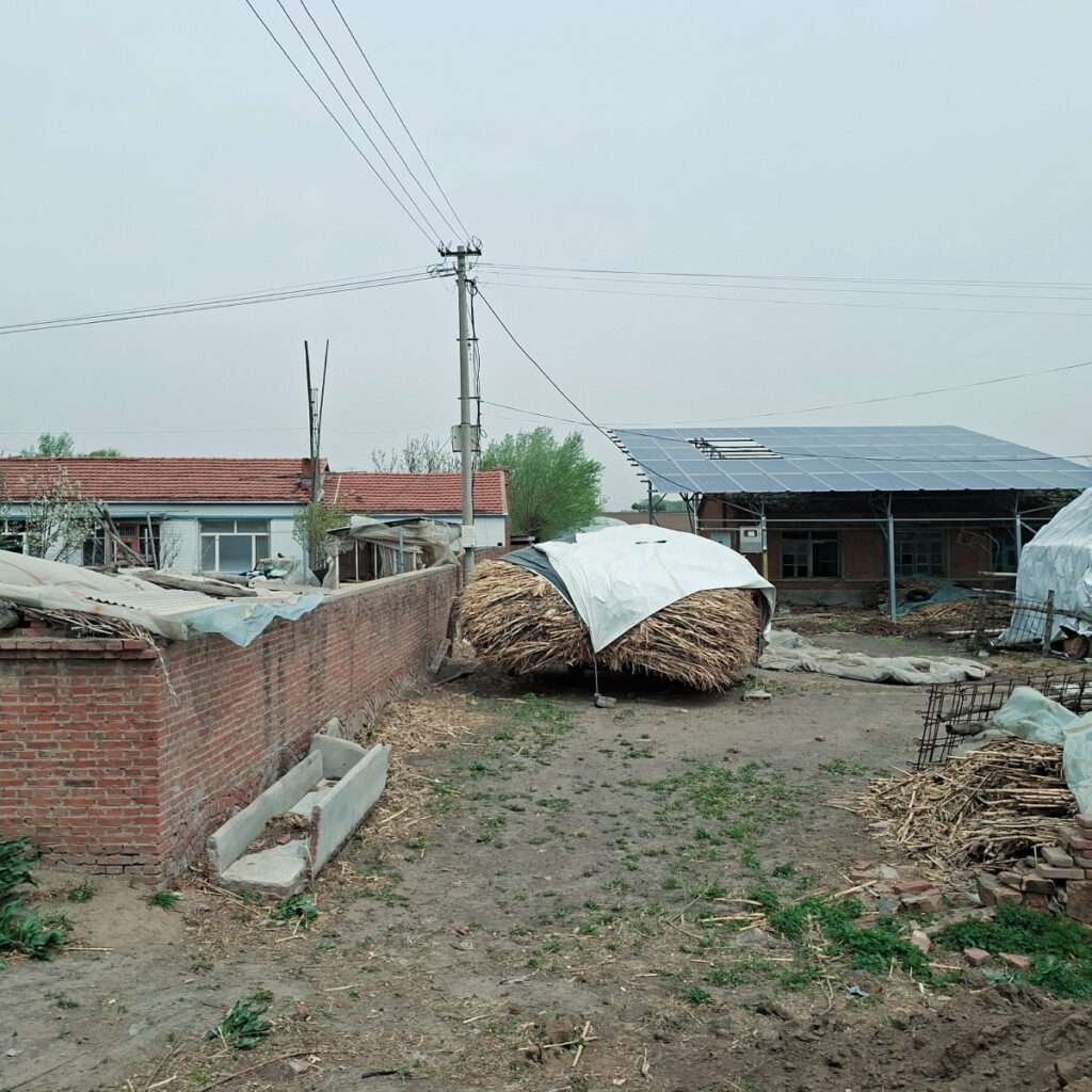 Rural yard with a hay bale under a white tarp beside a brick wall and a solar-panel shed underneath a cloudy sky.