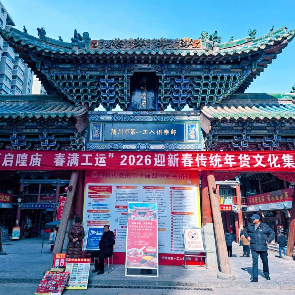 Ornate Chinese temple gate with blue-green tiles and decorative roof, a large red banner in front advertising a 2026 cultural event, and people selling posters nearby.