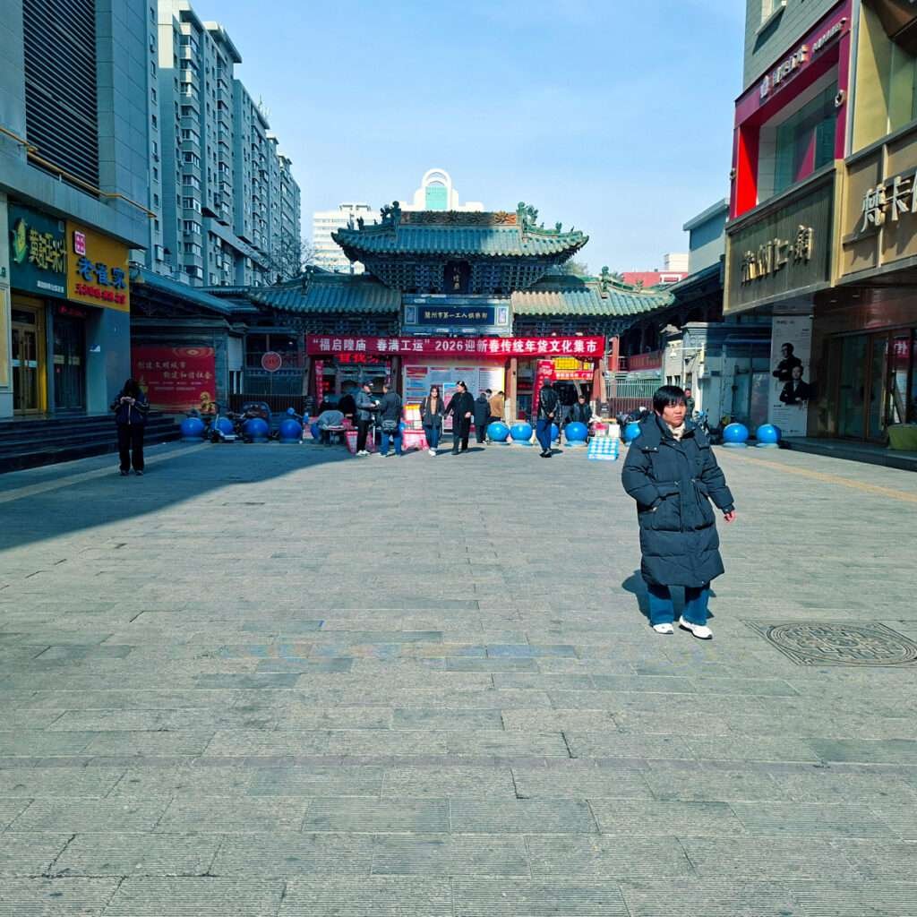 Wide pedestrian street toward a traditional Chinese gate with green tiled roof, red banners, and market stalls in a busy plaza.