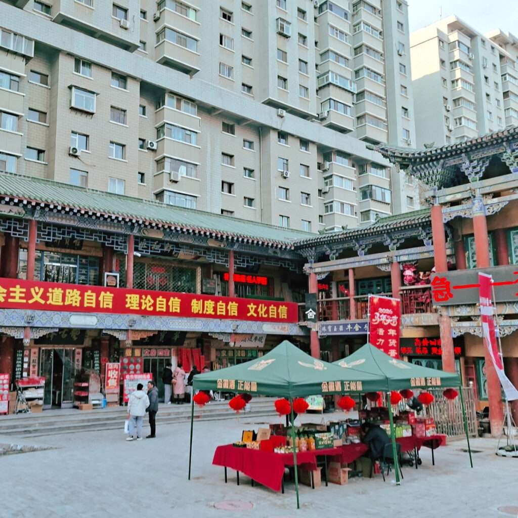 A bustling outdoor market in a Chinese plaza with red banners, vendors under green canopies, and high-rise apartment buildings behind.