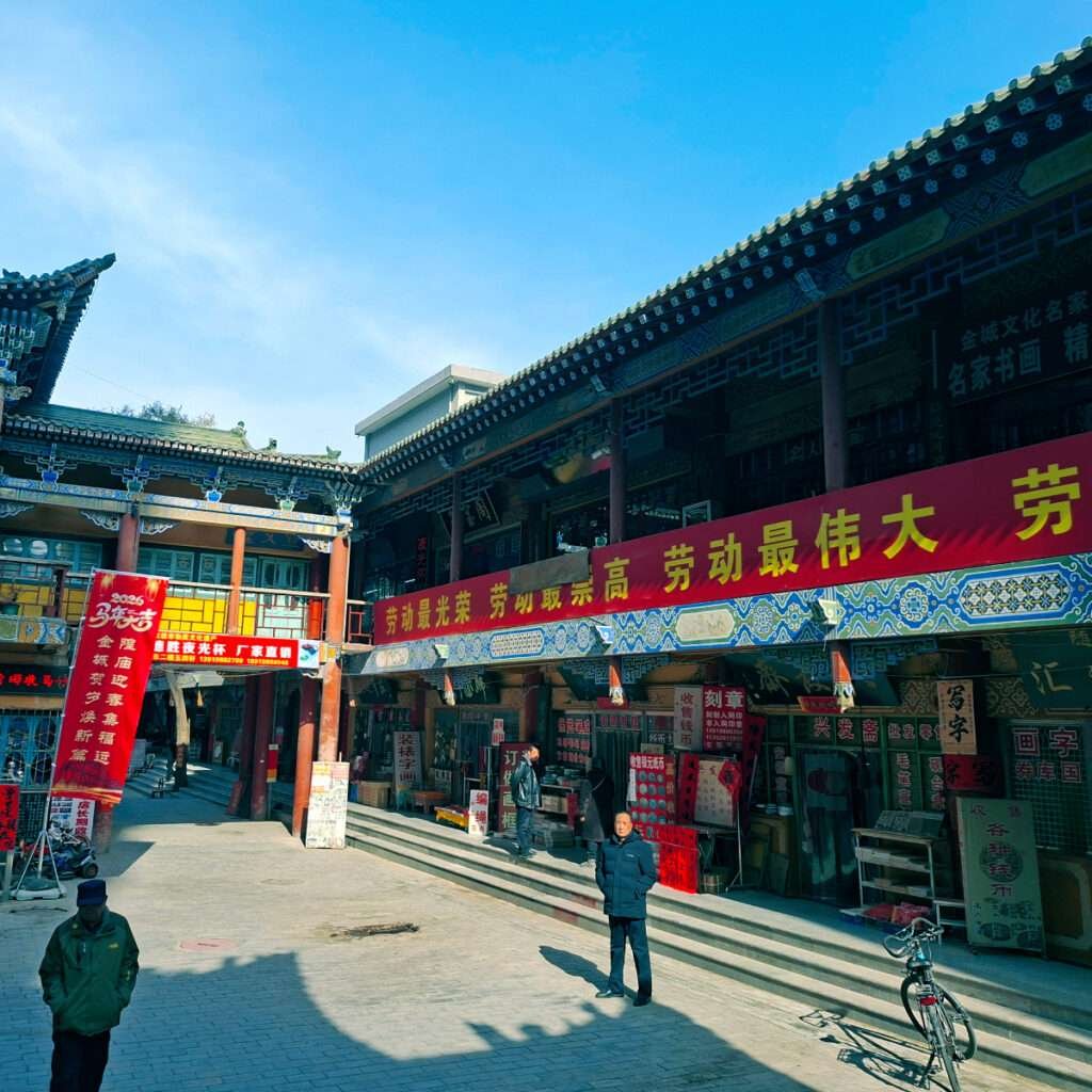 A covered market street with traditional Chinese architecture, red banners with yellow Chinese characters, and small shops along the walkway under a bright blue sky.