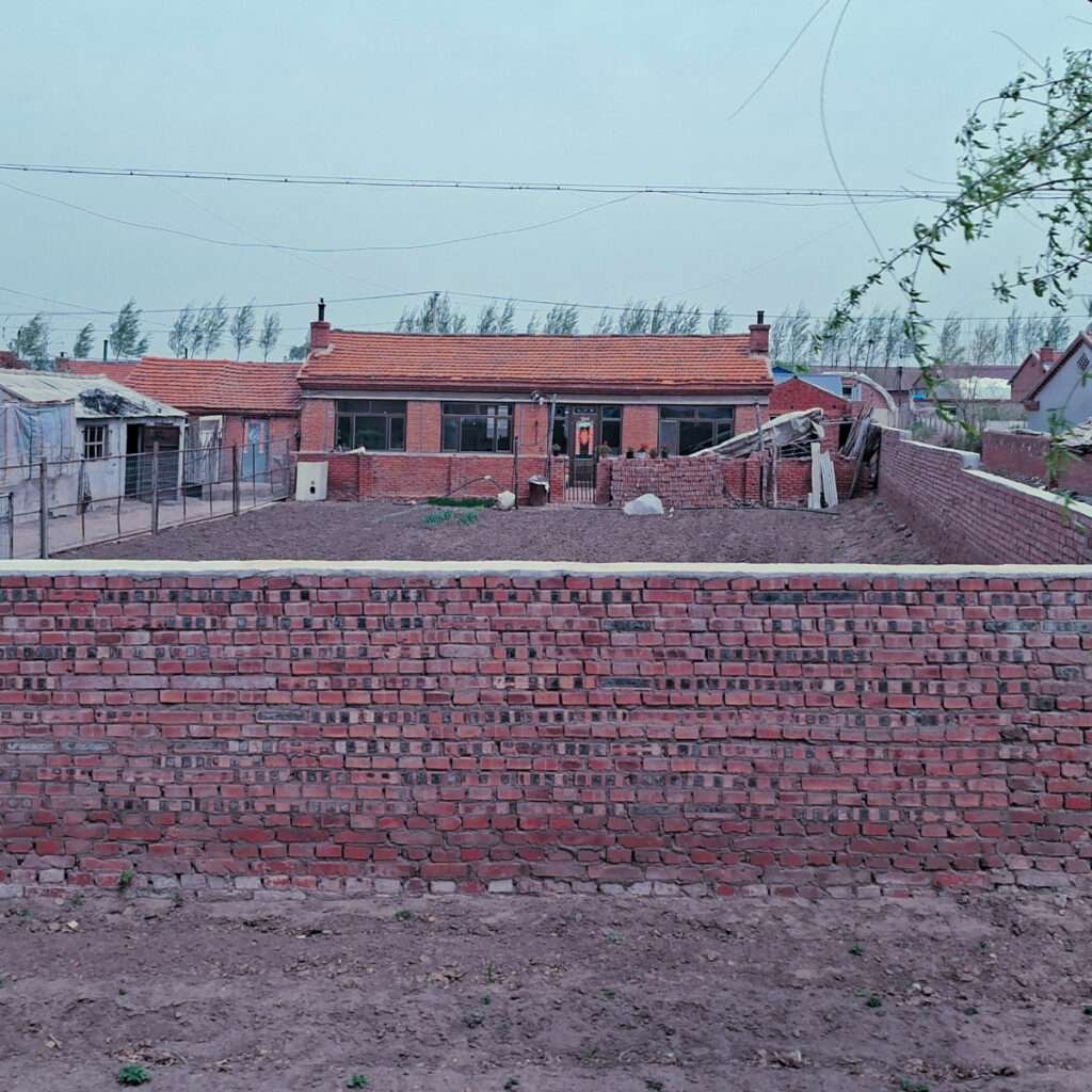 Front view of a single-story brick house with a red-tiled roof behind a long brick wall and a dirt yard.