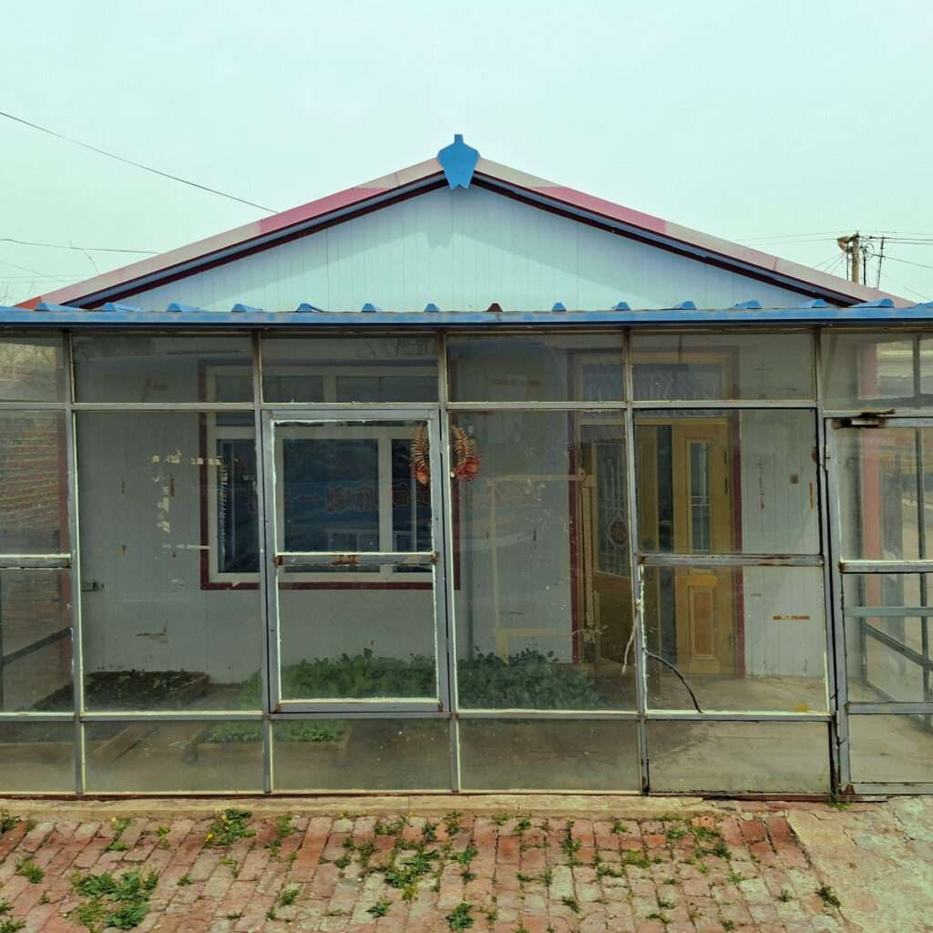 Front view of a glass-walled greenhouse with a blue roof and a door, weeds growing along a brick walkway in front.