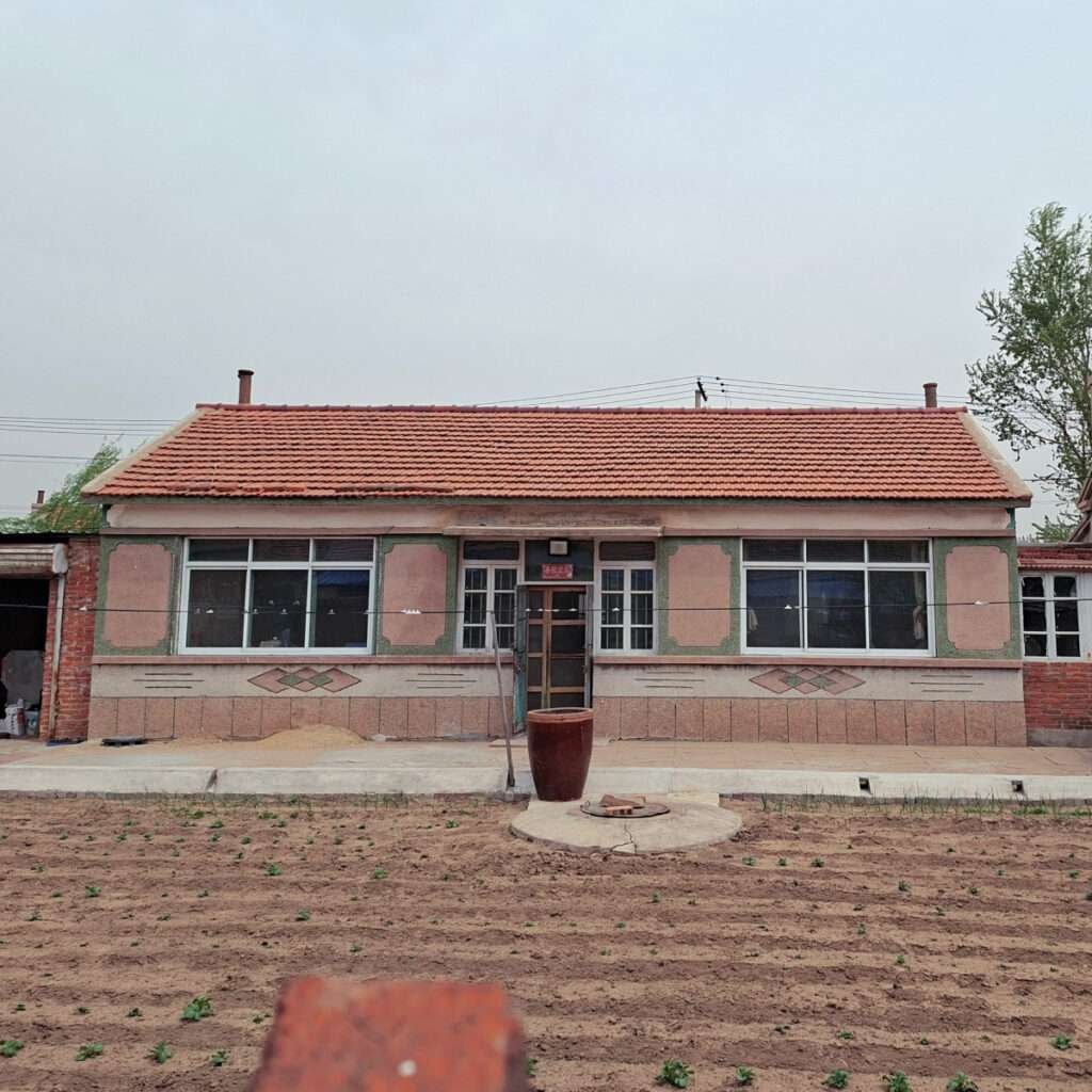 A single-story house with a red tiled roof and patterned exterior walls, a central door, and large windows; a tall ceramic pot sits on a circular concrete pad in front.
