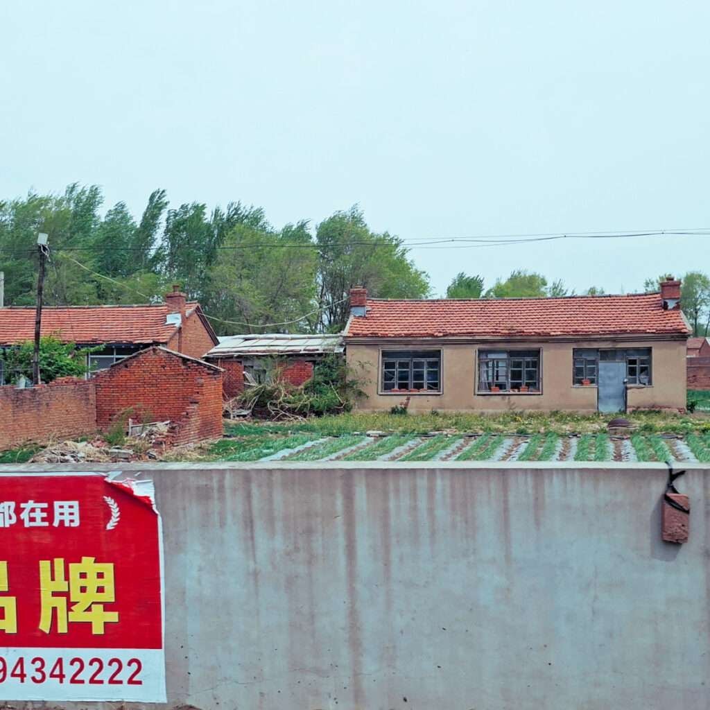Rural scene showing a row of brown houses with red-tiled roofs behind a concrete wall, with a large red Chinese ad banner in the foreground edge.