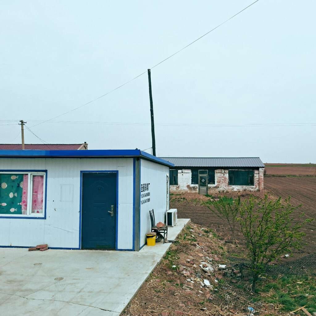 White and blue modular house with a blue door in a rural landscape; an older brick building and plowed fields in the background, with a yellow bucket and chair outside.