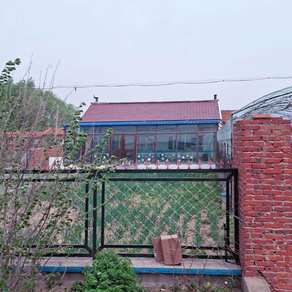 Brick gate and metal fence frame a backyard view of a glass-walled greenhouse-like structure with a red-tiled roof behind it.