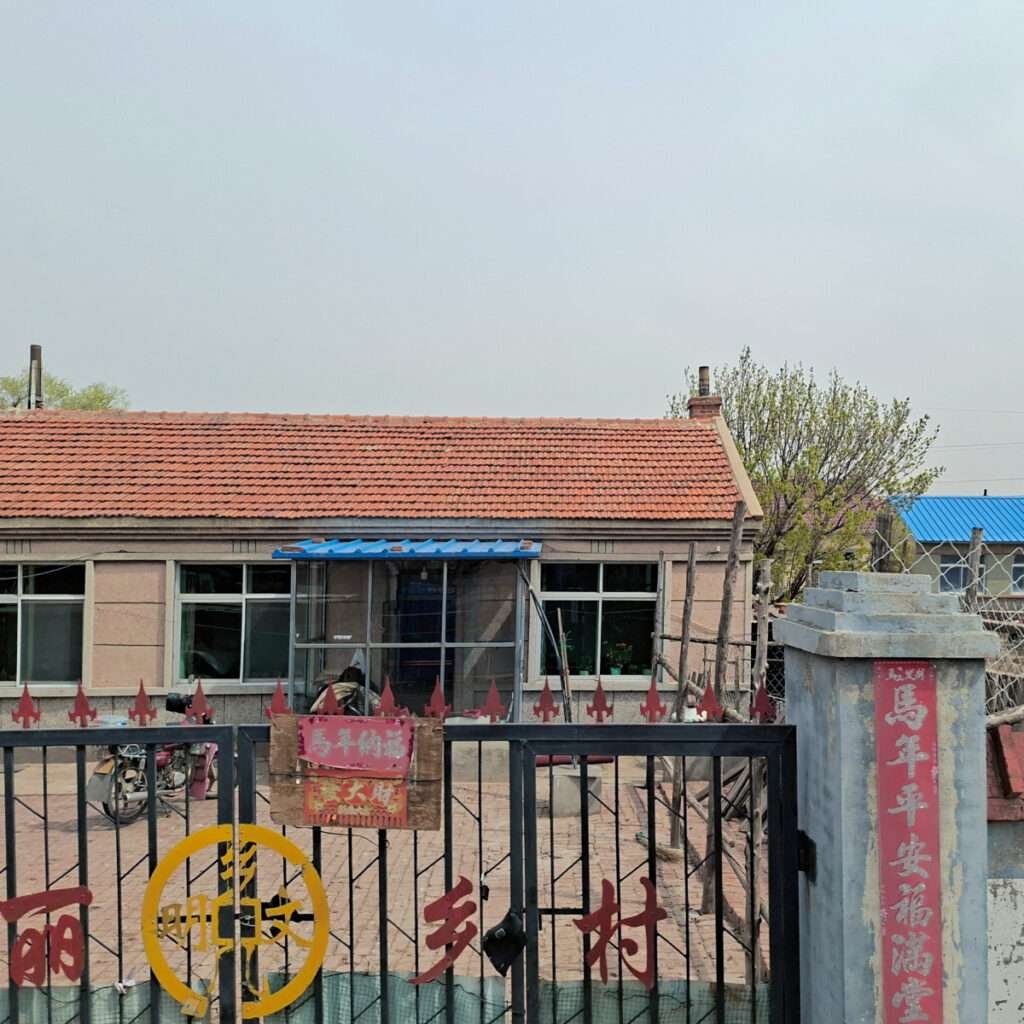 A small brick building with a red-tiled roof seen behind a black metal gate with red decorative panels and Chinese characters. A yellow circle sign is on the gate, and a blue awning covers the entrance. The scene is outdoors with a courtyard and sparse trees nearby.