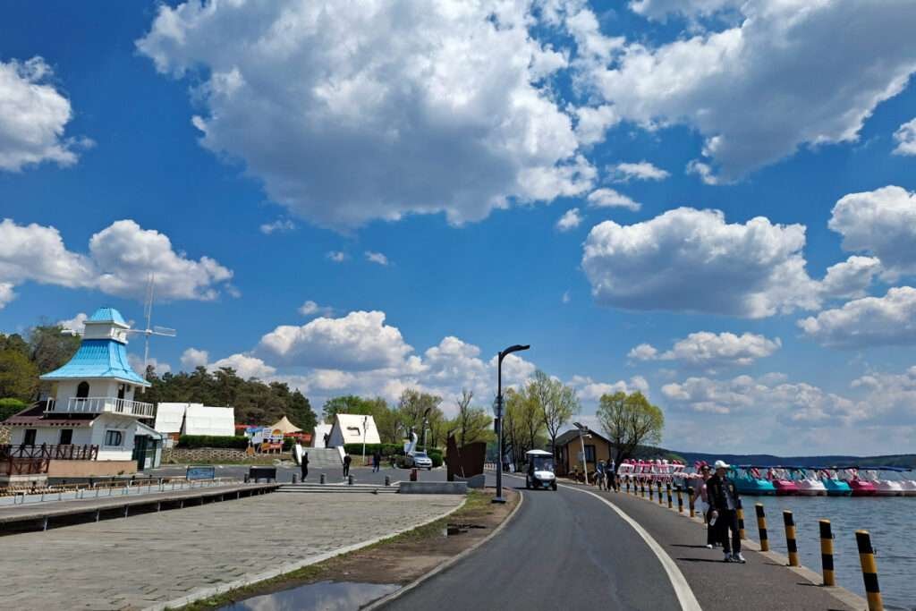 Sunny lakeside promenade with a blue-roofed white building, boats to the right, and people walking along the path.