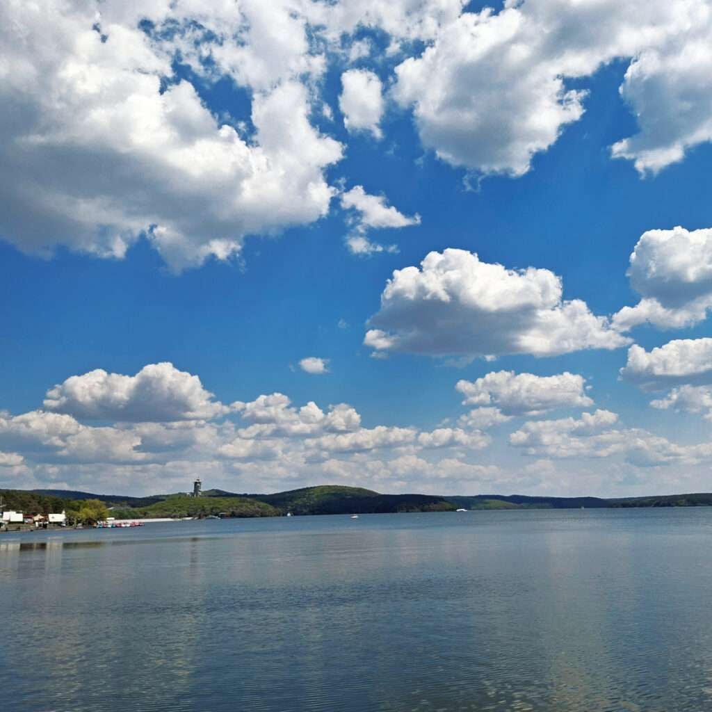 Calm lake extending to distant green hills with a lighthouse on the shore under a blue sky and white clouds.Note: avoid breaking text; use as single sentence.