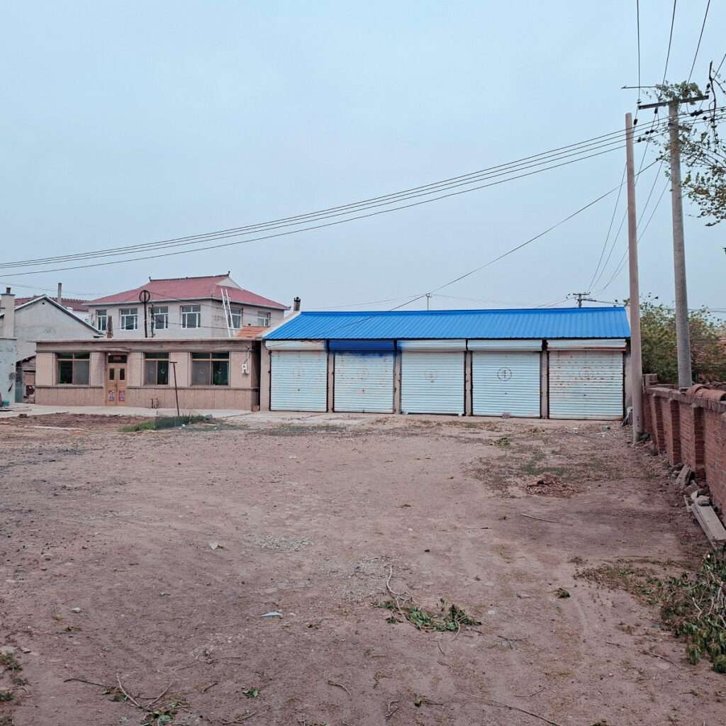 Row of closed storefronts with a blue metal roof, set in a dusty lot with power lines overhead.