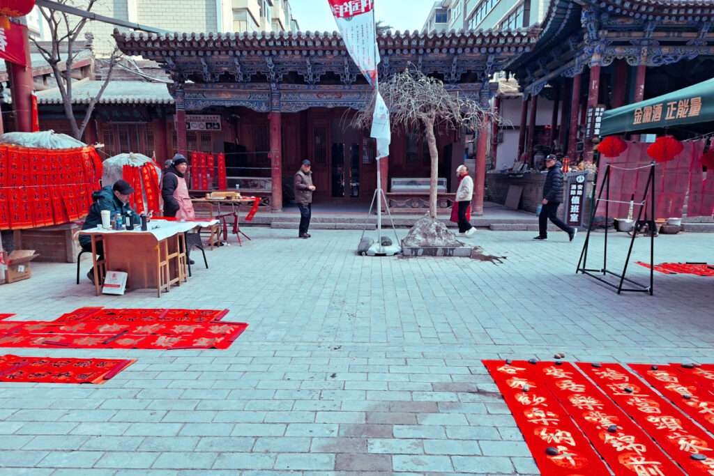 Temple courtyard with ornate wooden eaves, red banners on the ground, and people at a table setup under the gateway.