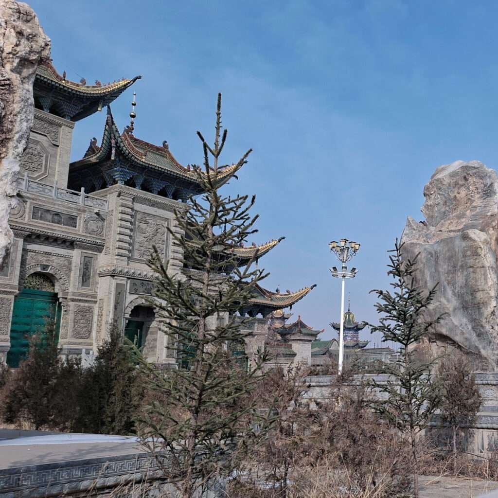 Ornate Chinese temple with carved stone walls, upturned tiled roofs, and evergreen trees against a blue sky.