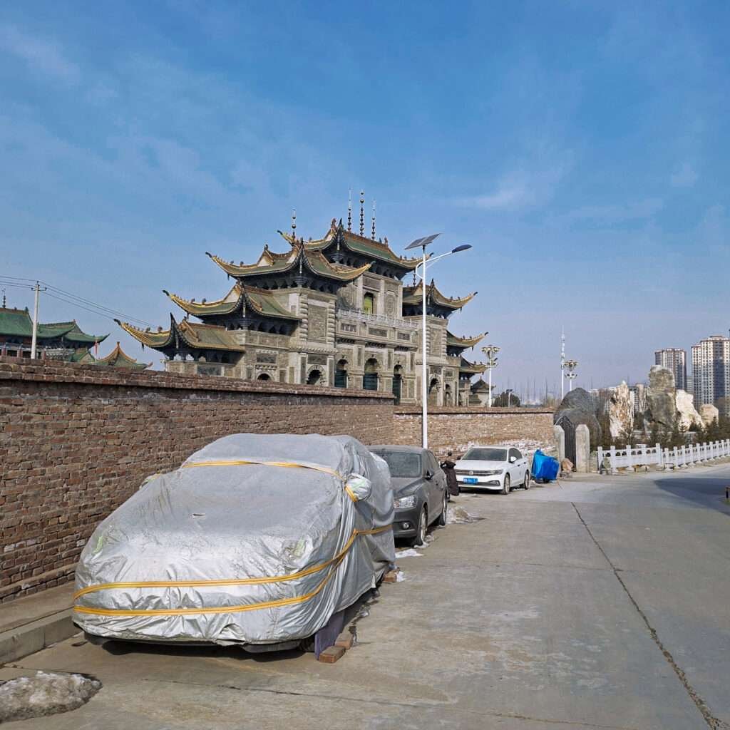 Stone Chinese temple behind a brick wall with multi-tier, upturned roofs and parked cars along the street behind it.