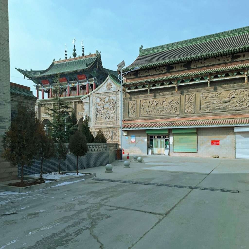 Ornate Chinese temple wall with carved stone panels and green tiled roof, adjacent to a paved courtyard and storefronts with closed shutters.
