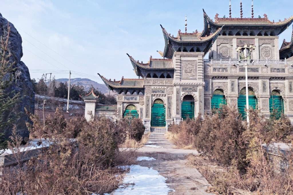 Ornate Chinese temple gate with green doors and carved stone walls amid barren winter vegetation and distant hills.