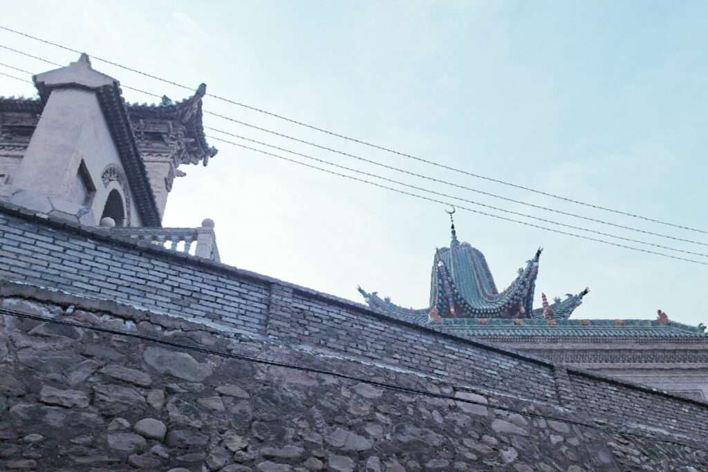 Traditional East Asian rooftops with ornate curved tiles and a stone wall in the foreground, under a clear blue sky.