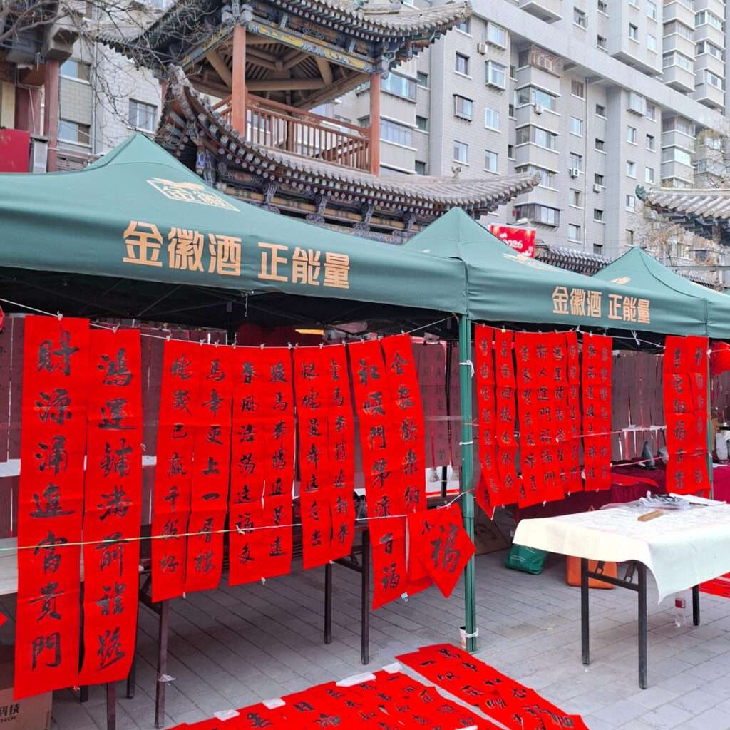 Green canopy with gold Chinese characters over a market stall; red vertical banners with black calligraphy for Lunar New Year decorations outside a building with traditional roofline.