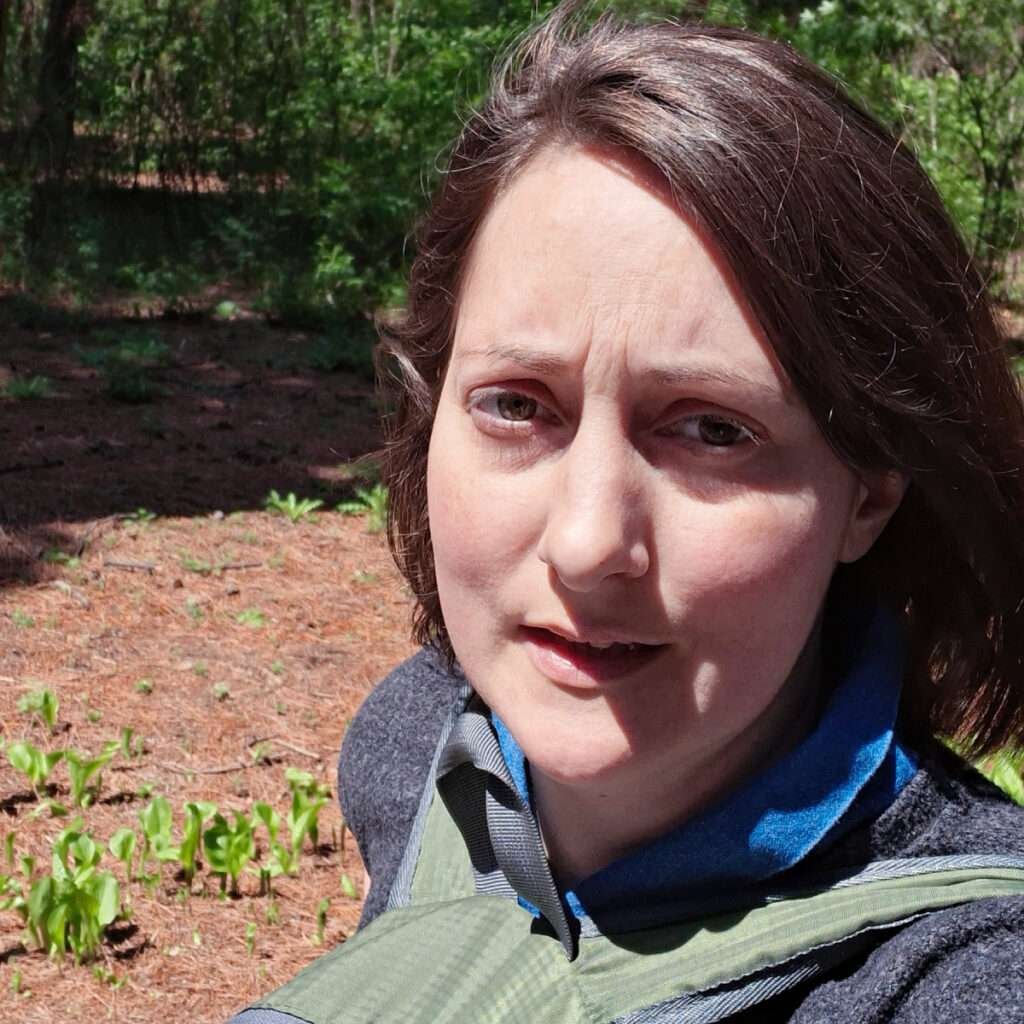 Close-up of a woman outdoors, sunlight on face, wearing a scarf and jacket in a forest setting.