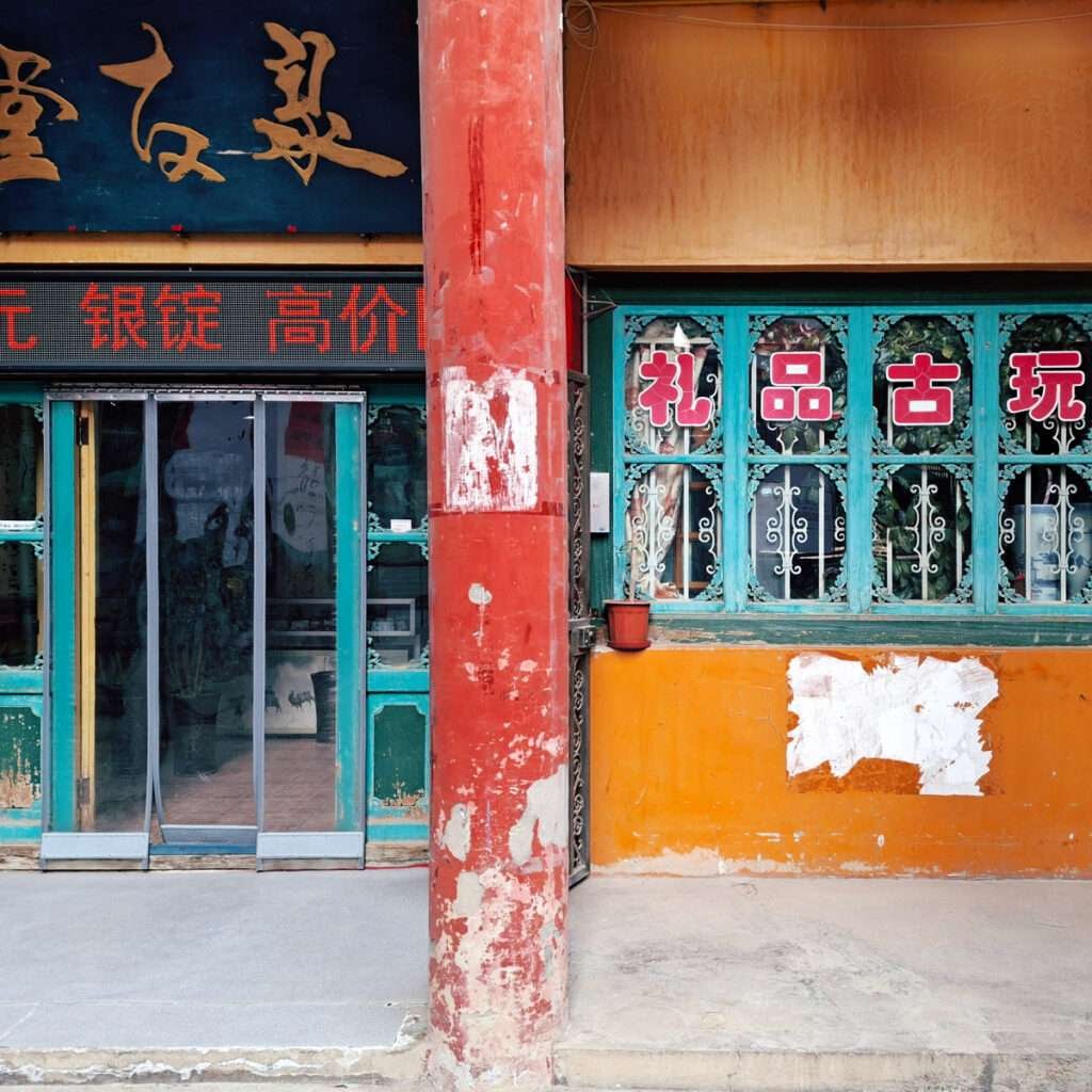 Colorful storefront with turquoise ornate window frames, orange wall, and a weathered red pillar in the foreground; Chinese characters on signs and a glass door storefront visible in the background.