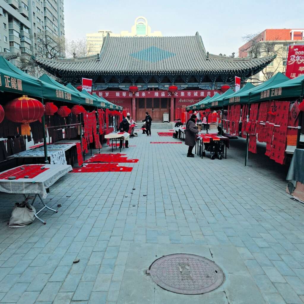 Outdoor market with green tents and red decorations in front of a traditional Chinese temple; red lanterns and banners abound, creating a festive scene.