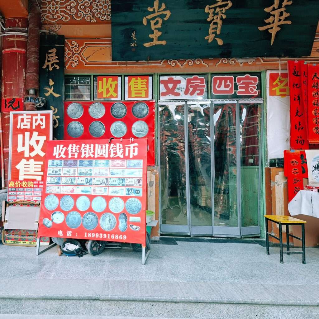 Shopfront with red signboards advertising coin collection and currency exchange; display board shows coins, Chinese characters on signs, and glass doors at entry.