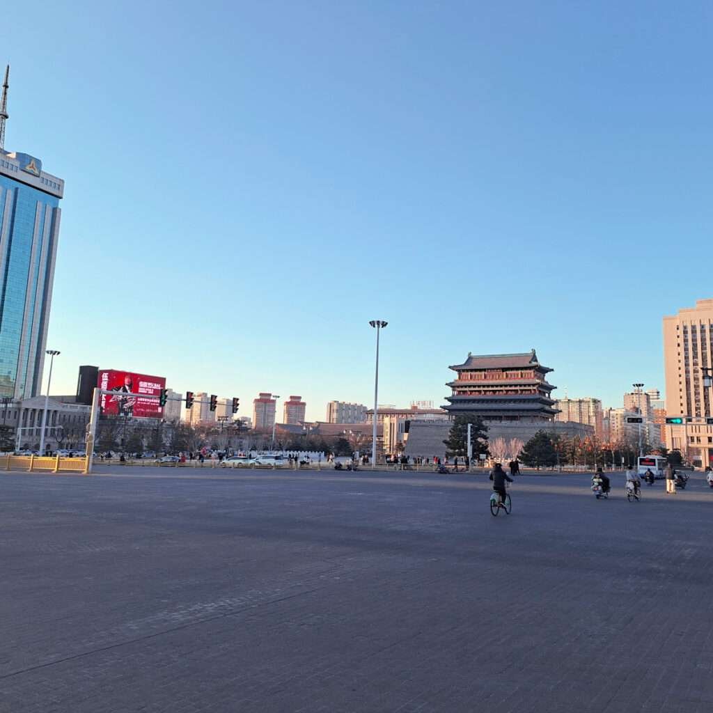 Public plaza with a historic Chinese gate and modern buildings on the horizon under a clear blue sky, people biking and walking around.
