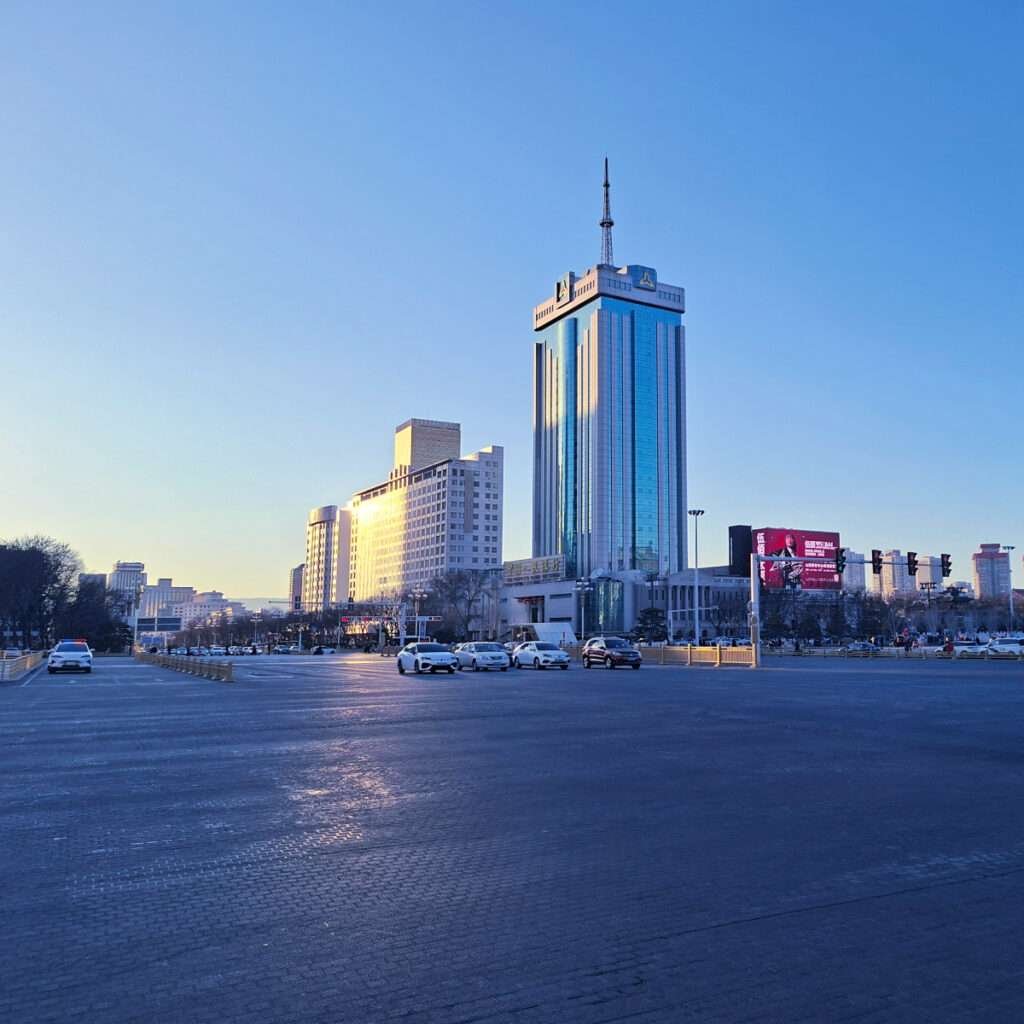 Tall blue-glass skyscraper with antenna, flanked by smaller buildings, along a wide city street at dusk with cars and a clear sky