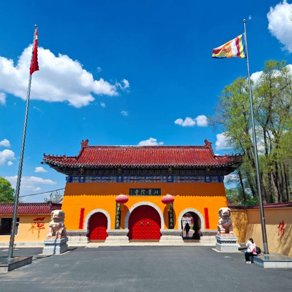 Chinese temple entrance with bright orange wall, red arched doors, and ornate roof, flanked by stone guardian lions against a blue sky.