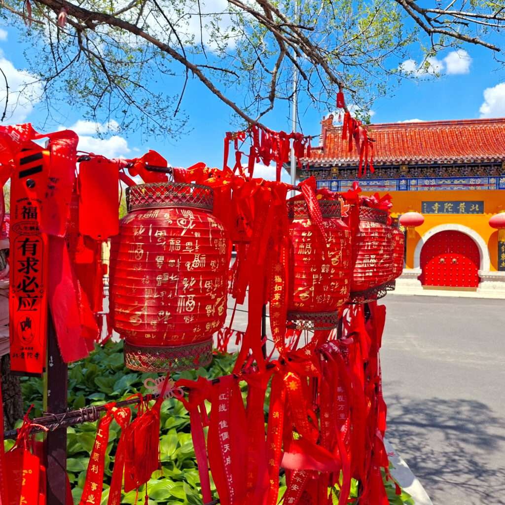 Red Chinese lanterns with gold inscriptions and red ribbons hanging in front of a temple entrance, blue sky above.