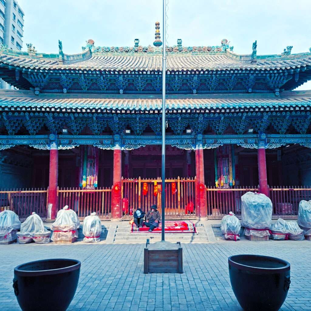 Two people sit on a red rug in front of a richly carved Chinese temple courtyard with blue beams and red columns under a decorative roof.”,