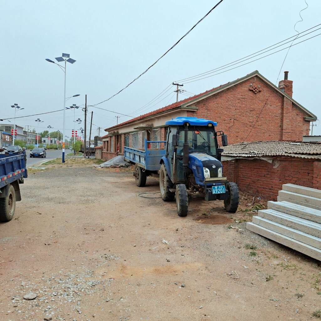 Blue tractor-trailer parked on a dirt road beside a brick building under construction with concrete slabs nearby.