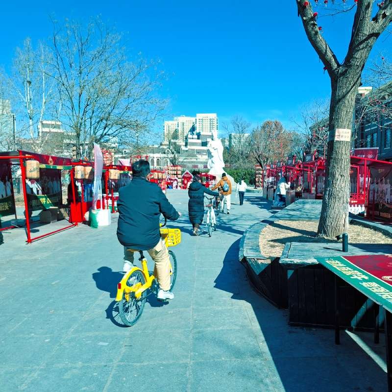 Sunny urban plaza with red market stalls lining the walkway; people ride bicycles and stroll under a clear blue sky.