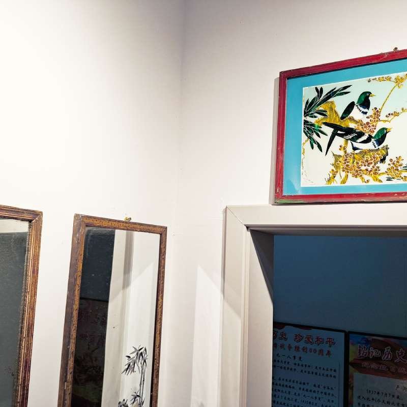 Corner scene with two wooden-framed mirrors leaning against a white wall; a colorful bird painting in a red frame sits above a doorway to a blue room.