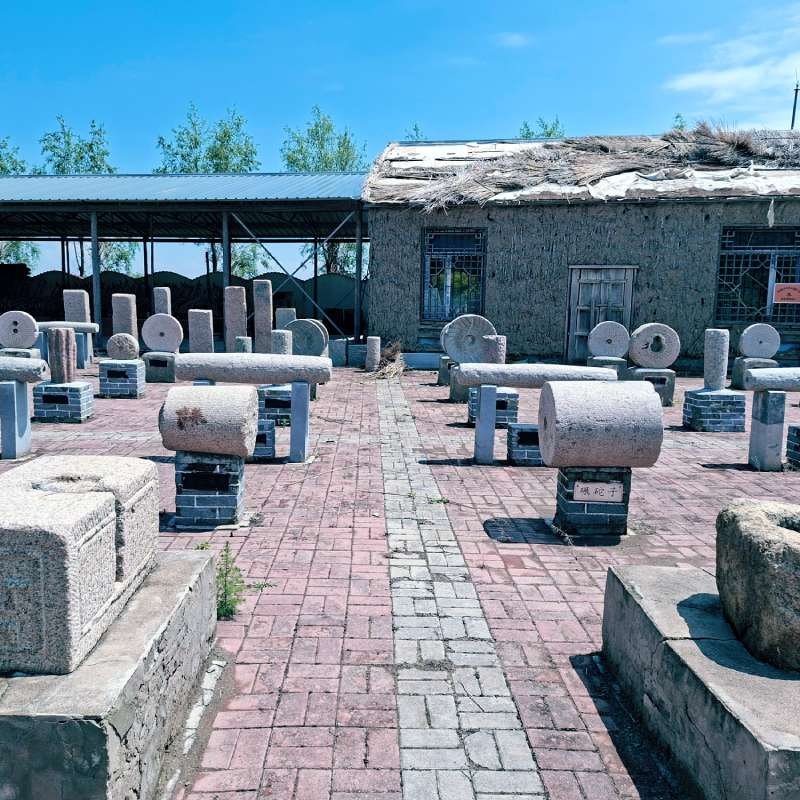 Outdoor sculpture yard with cylindrical and disc-shaped stone monuments on brick paving and a thatched-roof building in the background