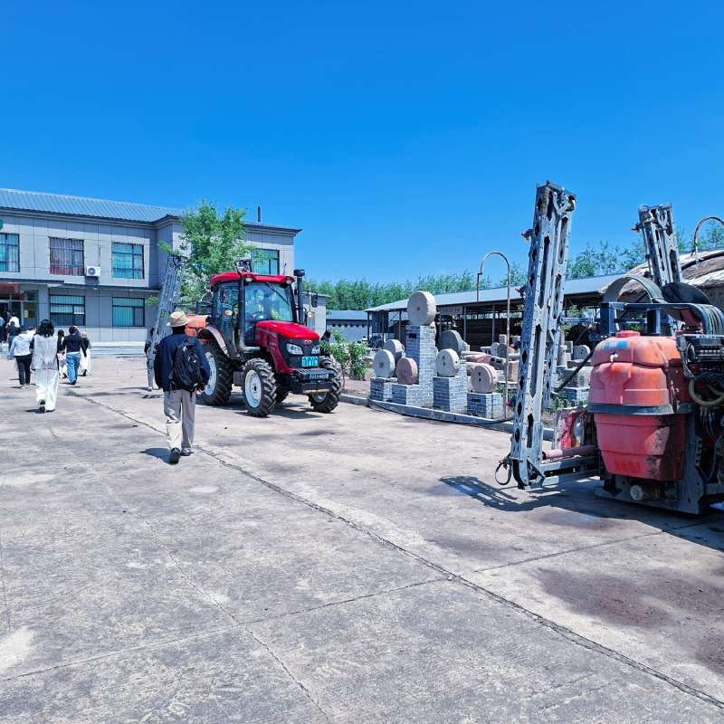 Red tractor parked in an outdoor construction yard beside stacks of circular stone pieces and other machinery, as people walk along the concrete lot.