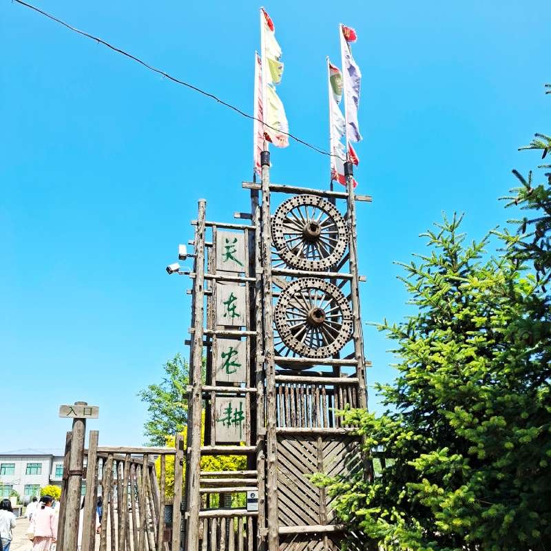 Tall wooden structure with two large wooden gears, vertical signs with green characters, and colorful flags against a blue sky; trees and people in the background.