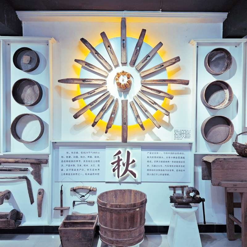 Exhibition wall with a sunburst of wooden blades around a central piece, illuminated from behind, and a large 秋 (autumn) character below, plus vintage farming tools and a wooden bucket in front.