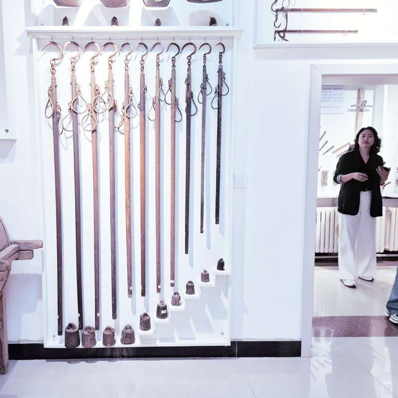Wall-length display of leather belts hanging from hooks in a bright showroom; a woman stands in a doorway to the right.